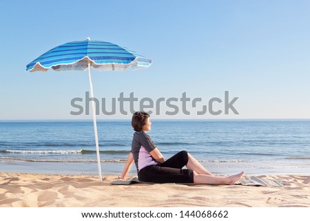 Middle-aged woman sitting on the beach carefree and relaxing. Under a beach umbrella.