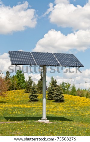 A photovoltaic solar panel array in a park with a blue sky and puffy white clouds in the background.