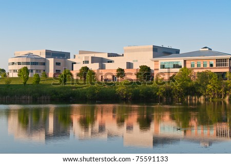 A community center building surrounded  by a park is shown reflected in the water of a large pond.