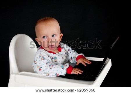 Baby sitting in the high chair, sticking out his tongue and using the laptop