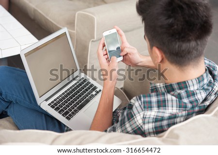 Businessman using smartphone and laptop writing on sofa in coffee shop