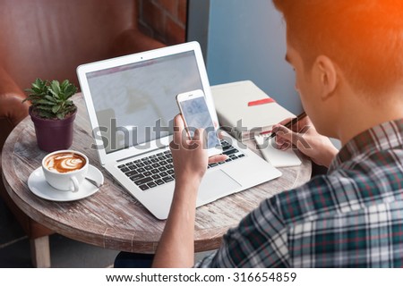 Businessman using smartphone and laptop writing on tablet on wooden table in coffee shop with a cup of coffee