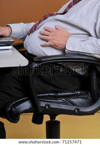 Middle aged man sitting in his office holding his large stomach while working at his desk. Sedentary men with heavy mid sections are more at risk for diabetes, heart attacks and strokes.