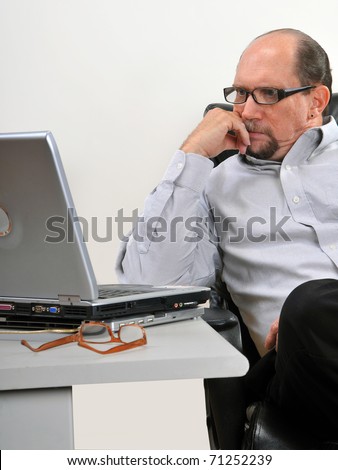 Middle aged man sitting at his desk looking at his computer in an office setting. He is concentrating on what he sees on his laptop.