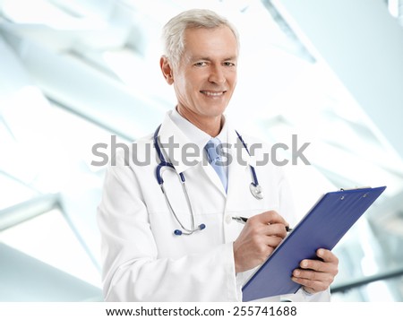 Portrait of happy mature medical doctor writing the result of medical test while standing at clinic.