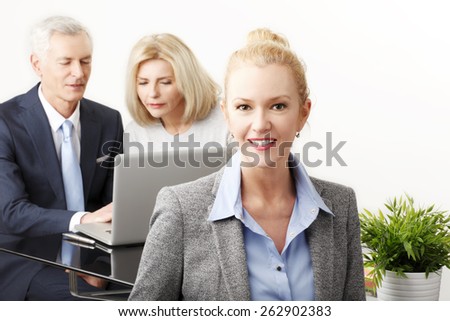 Portrait of executive business woman sitting at desk while business people working in background with laptop. Isolated on white background.