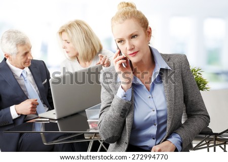 Attractive mature businesswoman sitting at office and making call. Sales woman and sales man working at background with laptop.