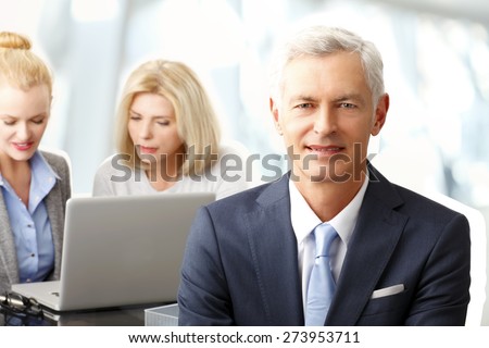 Portrait of executive senior businessman sitting at office and looking the camera. Busy sales women working on laptop at background.