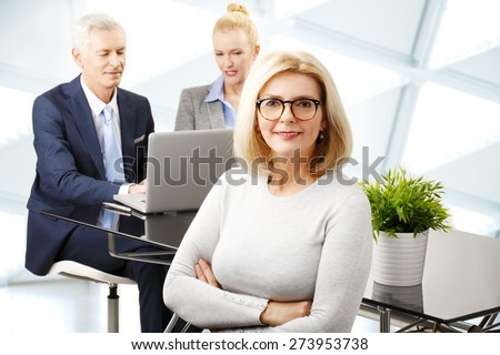 Image of executive businesswoman standing at office with arms crossed while financial assistant consulting at background and working at laptop.