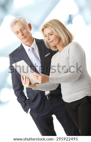 Portrait of middle age businesswoman standing at office while holding digital tablet in her hands. Senior businessman standing next to her and working together.