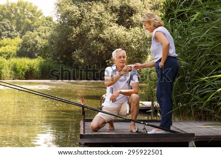 Happy senior couple relaxing at lakeside. Senior man squatting on the pier while her wife gives him a cup of coffee. Elderly people have fun.