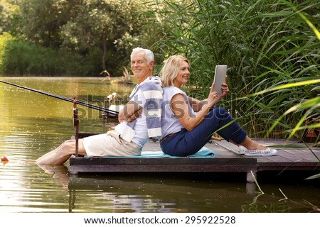 Portrait of elderly people sitting on pier at lakeside. Senior man fishing while his wife using digital tablet and surfing on internet.