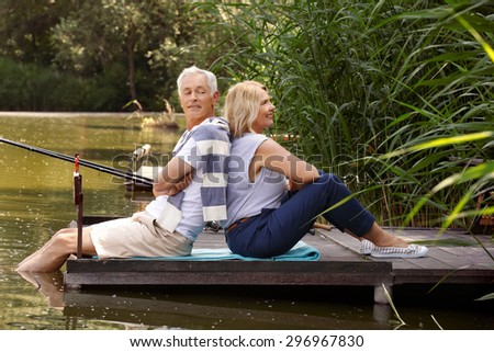 Elderly couple sit back to back each other on the pier. Happy senior people relaxing at the lakeside while they fishing.