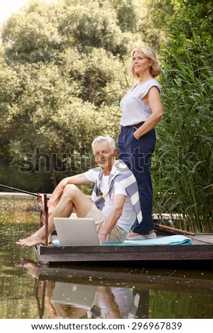 Portrait of senior couple fishing at lake. Elderly woman standing on the pier and senior man using laptop while surfing on the internet.