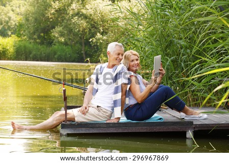 Portrait of elderly couple relaxing at nature. Senior woman and old man sitting back to back on the pier at lakeside while making self portrait with digital tablet.