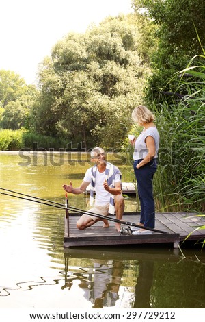 Portrait of senior couple fishing at the pier. Old man man squatting on the pier and shows to his wife how much fish caught. They are relaxing on th lakeside.
