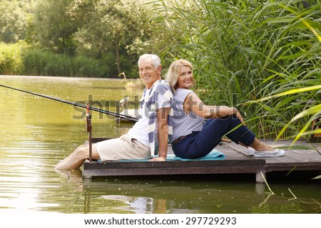 Elderly couple sit back to back each other on the pier. Happy senior people relaxing at the lakeside while they fishing.