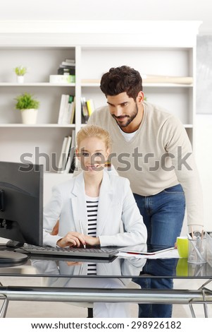 Portrait of young businesswoman sitting at desk in front of computer while smiling creative director standing next to her and consulting. Business people working together.