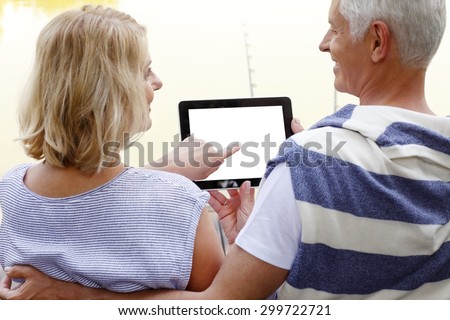 Close-up portrait of senior people with digital tablet sitting at the lakeside and surfing on internet. Elderly woman holding digital tablet displaying a white screen while old man hugging his wife.