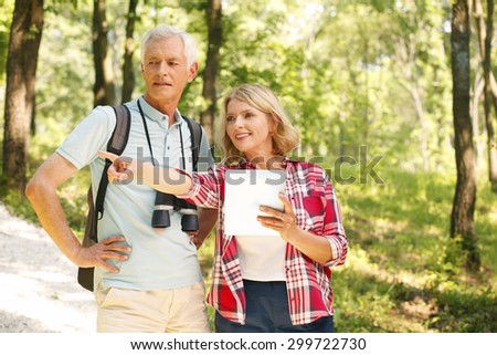 Portrait of senior couple take an excursion. Elderly man with binoculars standing at forest while senior woman holding hand digital tablet while her finger shows the route.