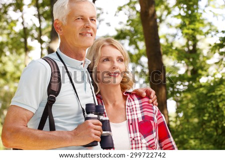 Portrait of happy senior people enjoying a walk in the forest together. Elderly man holding hands binoculars while senior woman smiling and standing next to him.