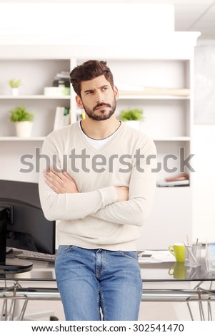 Portrait of businessman standing at office in front of computer and thinking. Young creative wearing casual clothing.