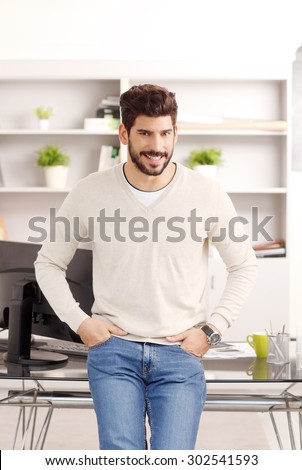 Portrait of young businessman standing at office desk in front of his personal computer. Creative professional wearing casual clothing and looking at camera while smiling.