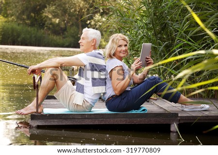 Portrait of senior couple chilling out at lake shore. Elderly man sitting on the pier and fishing while senior woman sitting with her back to him and holding in hand digital tablet.