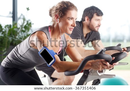 Close-up portrait of gym members participating in a spinning class while training together at fitness center. Sporty woman listening music at her mobile phone.