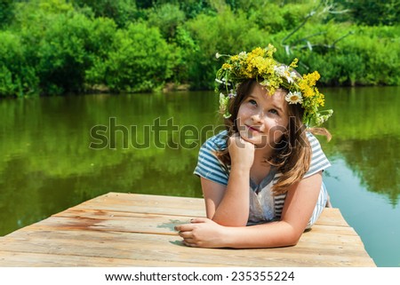 Beautiful little girl in a wreath of wild flowers lying on a wooden bridge on the river in summer