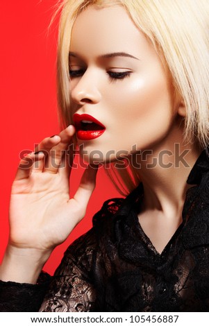 High fashion portrait of beautiful sexual woman in dynamic model pose, black lacy shirt on bright red background. Red lips make-up, straight blond hair