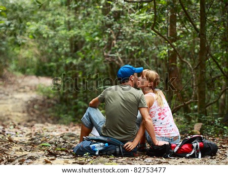 Two young kissing people sitting on the backpack in the forest