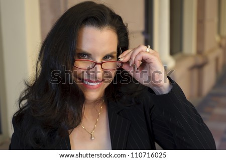 Portrait of an attractive middle aged woman smiling and peeking up through her red reading glasses. Shot on location with a business offices in the background.