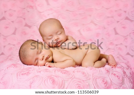 Fraternal twin newborn baby girls sleeping on pink, three dimensional rose fabric. One baby is lying on her stomach and the other is propped on top of her sister.