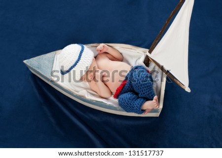One week old newborn baby boy wearing a white and blue sailor hat. It is an overhead view of him sleeping on his back in a tiny sailboat. Shot in the studio on blue velvet.