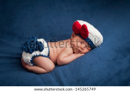 Eight day old newborn baby girl wearing a white and blue sailor costume. She is sleeping contentedly on her stomach. Shot in the studio on navy blue velvet.
