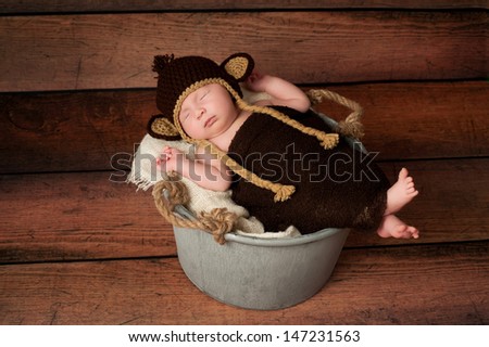 A newborn baby wearing a crocheted monkey costume and sleeping in a galvanized bucket. Shot in the studio with a rustic wood background.