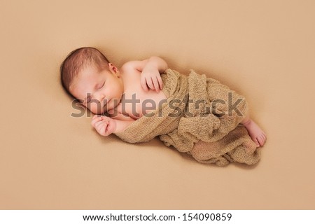 A 3 week old newborn baby boy wrapped in gauzy beige fabric and sleeping on a beige fleece blanket.