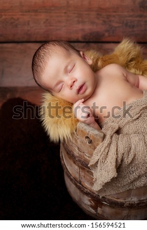 A newborn baby boy sleeping in an antique wooden well bucket. Shot in the studio with a sheepskin rug and rustic wood background.