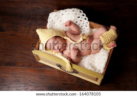 Portrait of a 2 week old sleeping newborn baby girl. She is wearing a yellow crocheted pixie hat and leg warmers and sleeping in a vintage drawer. Shot from overhead on a dark wood background.