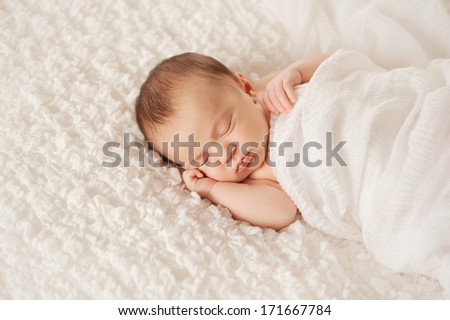 Head and shoulders shot of a sleeping two week old newborn baby wrapped in white gauzy fabric and sleeping on a white billowy blanket.