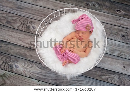 Newborn baby girl sleeping in a white, wire basket lined with white faux fur. She is wearing a pink and white, vintage inspired, sleeping cap with matching legwarmers.