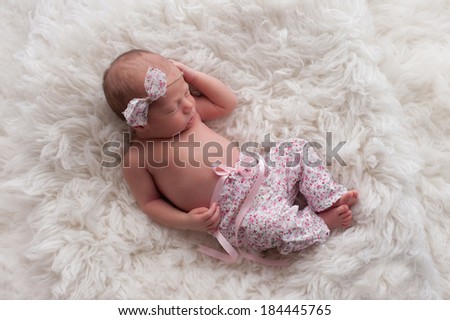 Portrait of a ten day old sleeping newborn girl wearing white and pink floral pants with a matching headband. She is sleeping on a white Flokati (sheepskin) rug.