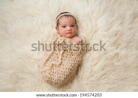 An alert 7 day old, Hispanic baby girl with a displeased look on her face. She is wrapped in a crocheted wool pouch and looking at the camera. Shot in the studio on a beige flokati (sheepskin) rug.