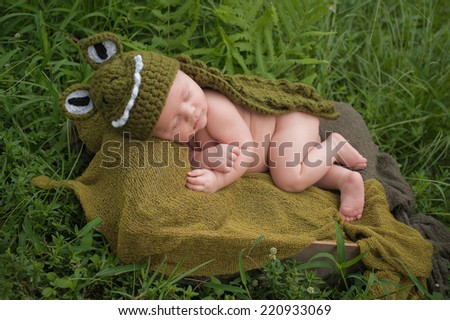 A three month old baby boy wearing a green alligator costume. He is sleeping in a crate that\'s placed outside in a grassy area.