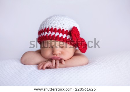 Portrait of a seven day old newborn baby girl. She is wearing a white and red crocheted hat and is sleeping on white, textured material.