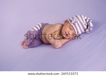 Smiling eight day old newborn baby girl sleeping peacefully. She is wearing a striped, lilac and white, upcycled sleeping cap and pant set.