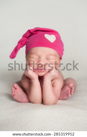 A portrait of a beautiful eleven day old baby girl wearing an up-cycled pink top knot cap with heart detail. She is sleeping on a beige blanket and posed with her chin in her hands.