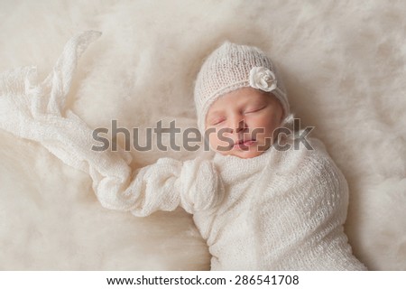 A portrait of a beautiful, seven day old newborn baby girl wearing a white, knitted, mohair bonnet. She is swaddled with a gauzy, white fabric and sleeping on cream colored wool batting.