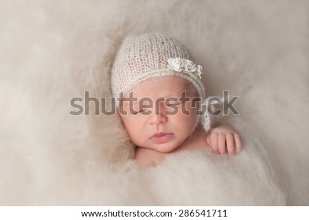A portrait of a beautiful seven day old newborn baby girl wearing a white, knitted, mohair bonnet and rose headband. She is sleeping in a bed of cream colored wool batting.